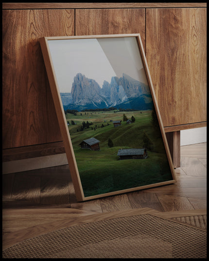 Ein gerahmtes Poster von einer grünen Alm mit kleinen Hütten und den Dolomiten im Hintergrund, an einer Holzkommode angelehnt und stehend auf dem Holzboden