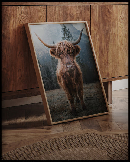 Ein gerahmtes Poster von einem Highland-Rind, das in einer Gebirgslandschaft steht, an einer Holzkommode angelehnt und stehend auf dem Holzboden