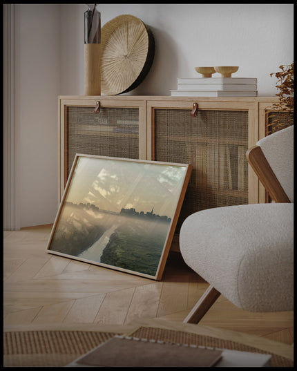 Ein gerahmtes Poster von einer nebligen Landschaft in den Niederlanden, mit einem Wasserkanal und einer Kirche im Hintergrund, an einer Holzkommode angelehnt und stehend auf dem Holzboden