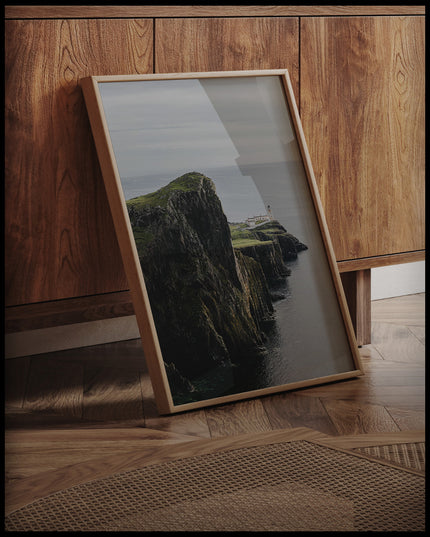 Ein gerahmtes Poster von einer kargen Klippe in Schottland mit einem Leuchtturm, an einer Holzkommode angelehnt und stehend auf dem Holzboden