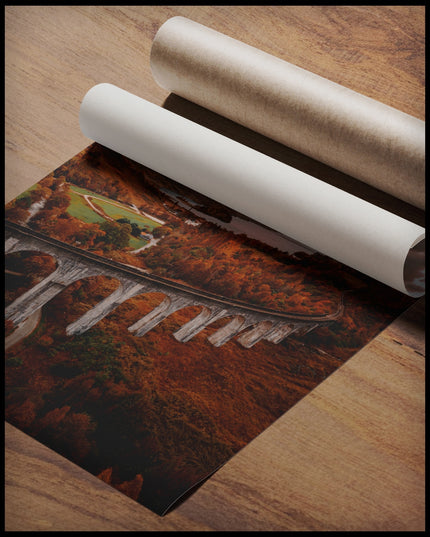 Ein Poster von dem Glenfinnan-Viadukt in Schottland, umgeben von herbstlicher Landschaft und Bergen im Hintergrund, gerollt auf einer Holzoberfläche liegend