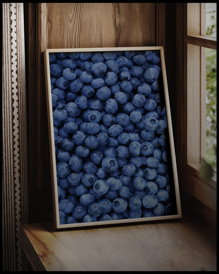 Ein gerahmtes Poster von einer Fülle von frischen Blaubeeren, die das gesamte Bild bedecken, vor einem Fenster an die Wand angelehnt