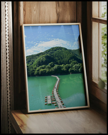 Ein gerahmtes Poster von einer schwimmenden Brücke auf einem grünlichen See, die in einen Wald hinein führt, vor einem Fenster an die Wand angelehnt