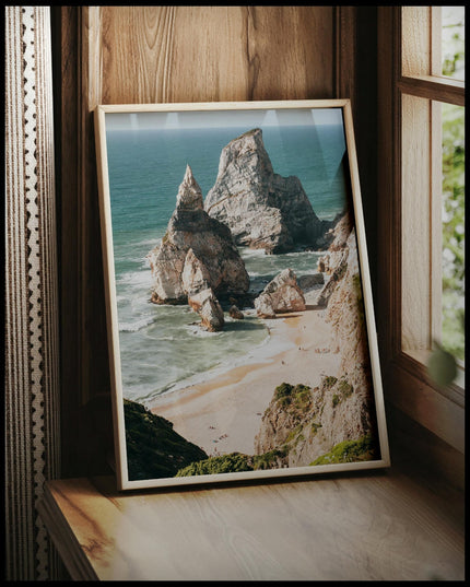 Ein gerahmtes Poster von hohen Felsformationen im Meer an einem Strand, vor einem Fenster an die Wand angelehnt