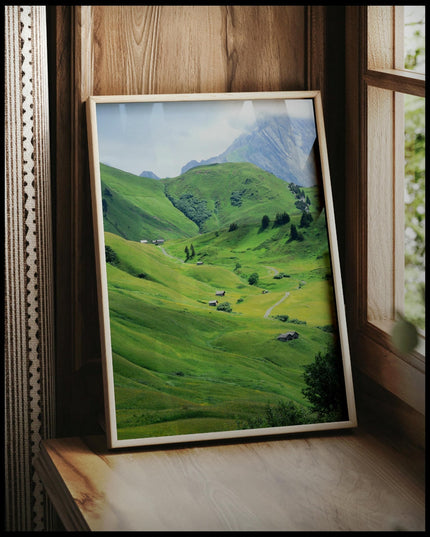 Ein gerahmtes Poster von einer grünen Berglandschaft mit einzelnen Bäumen und Holzhütten, vor einem Fenster an die Wand angelehnt